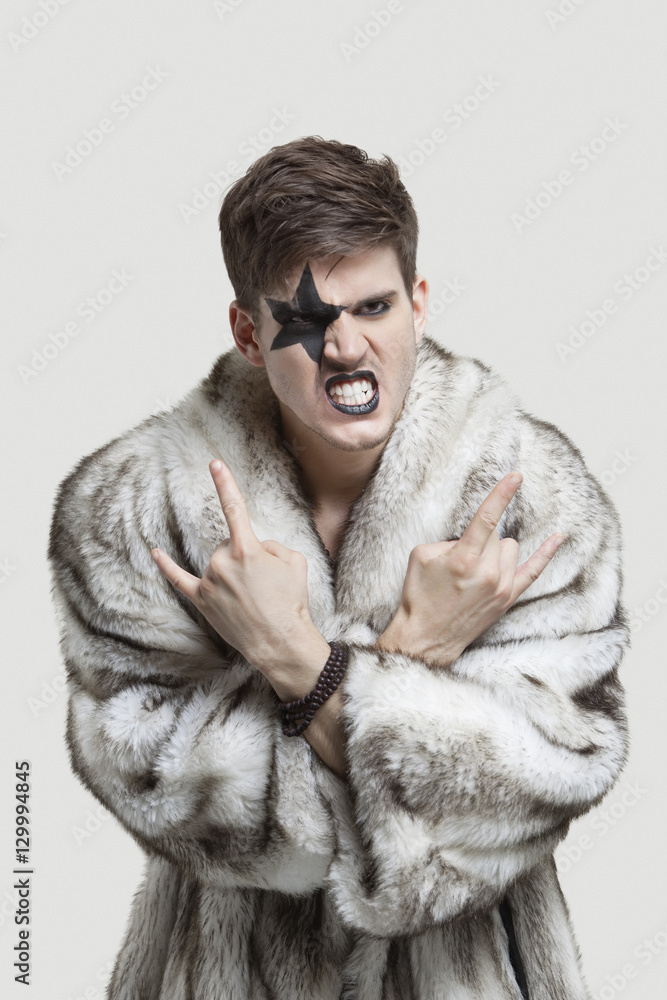 Portrait of frustrated young man in fur coat clenching teeth and making rebellious gesture against gray background Portrait of frustrated young man in fur coat clenching teeth and making rebellious gesture against gray background
