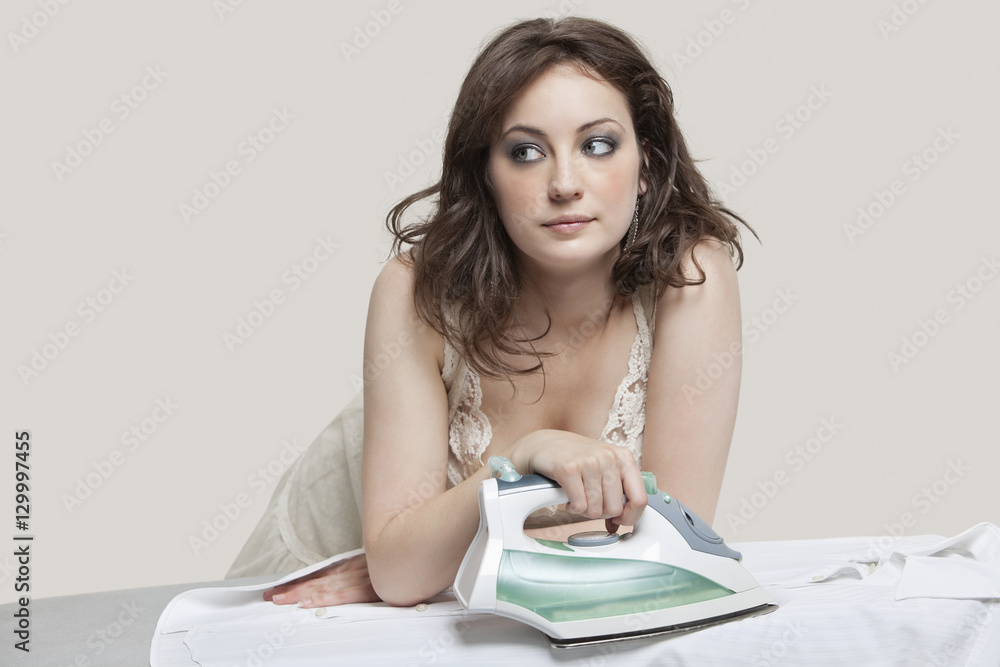 Young woman looking away while ironing shirt over gray background Young woman looking away while ironing shirt over gray background