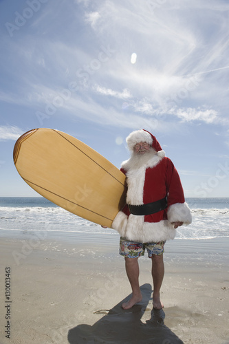 Photography Portrait of happy man dressed in Santa Claus outfit holding surfboard on beach