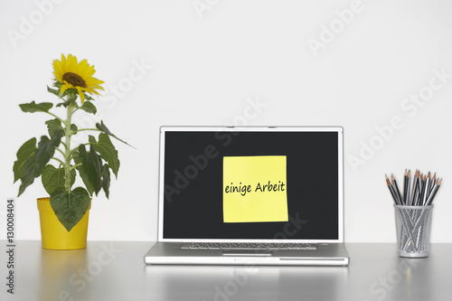 Sunflower plant on desk and sticky notepaper on laptop screen with  einige Arbeit  written on it in German