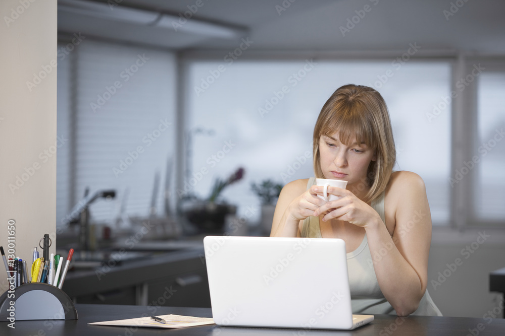Unhappy young woman holding teacup while looking at laptop in kitchen Unhappy young woman holding teacup while looking at laptop in kitchen