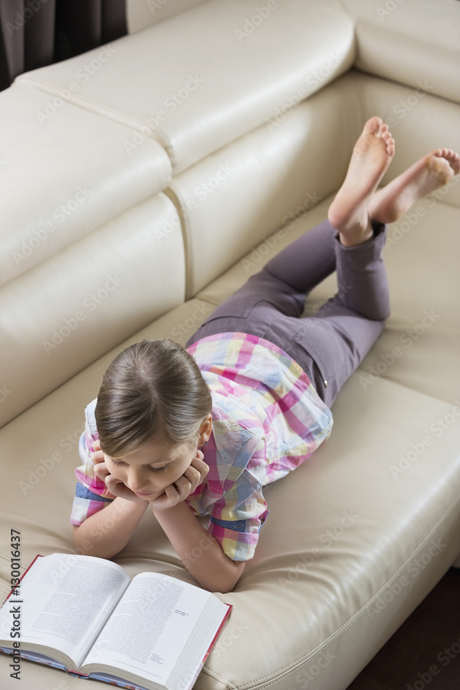 High angle view of girl reading book while lying on sofa at home High angle view of girl reading book while lying on sofa at home