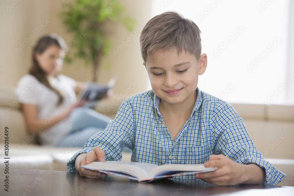Cute boy reading book with mother in background at home Cute boy reading book with mother in background at home