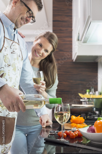 Happy man pouring white wine in glass while cooking with woman at kitchen