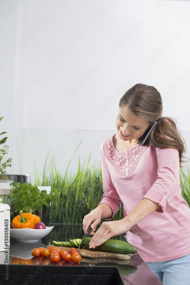 Smiling woman using cell phone while cutting cucumber at kitchen counter Smiling woman using cell phone while cutting cucumber at kitchen counter