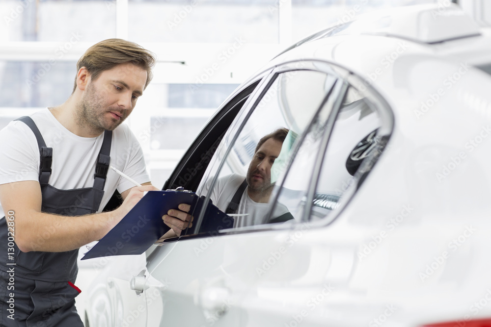 Automobile mechanic writing on clipboard while examining car in repair shop Automobile mechanic writing on clipboard while examining car in repair shop