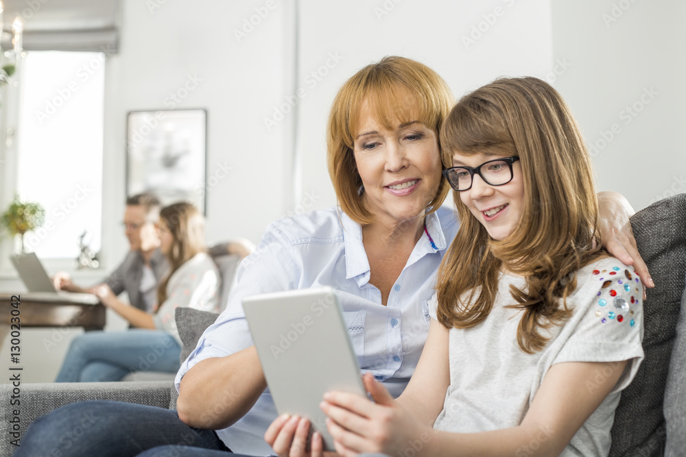 Loving mother and daughter using tablet PC with family sitting in background at home Loving mother and daughter using tablet PC with family sitting in background at home
