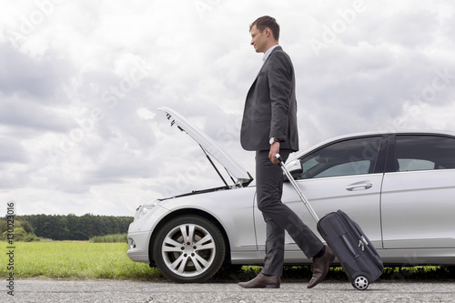 Full length side view of young businessman with luggage walking by broken down car at countryside © moodboard