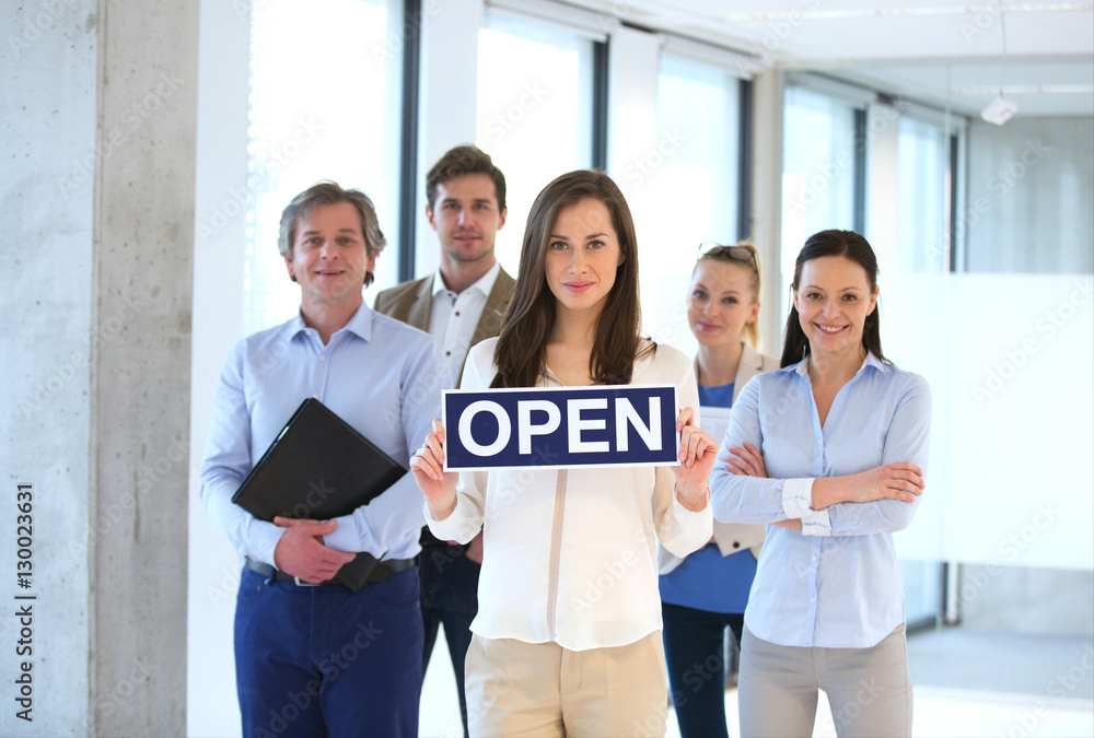 Portrait of young businesswoman holding open sign with team behind at office Portrait of young businesswoman holding open sign with team behind at office
