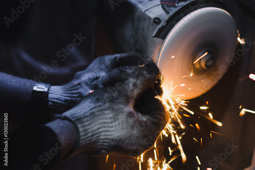 Factory worker at workshop grinding steel rod with abrasive disc and flying sparks