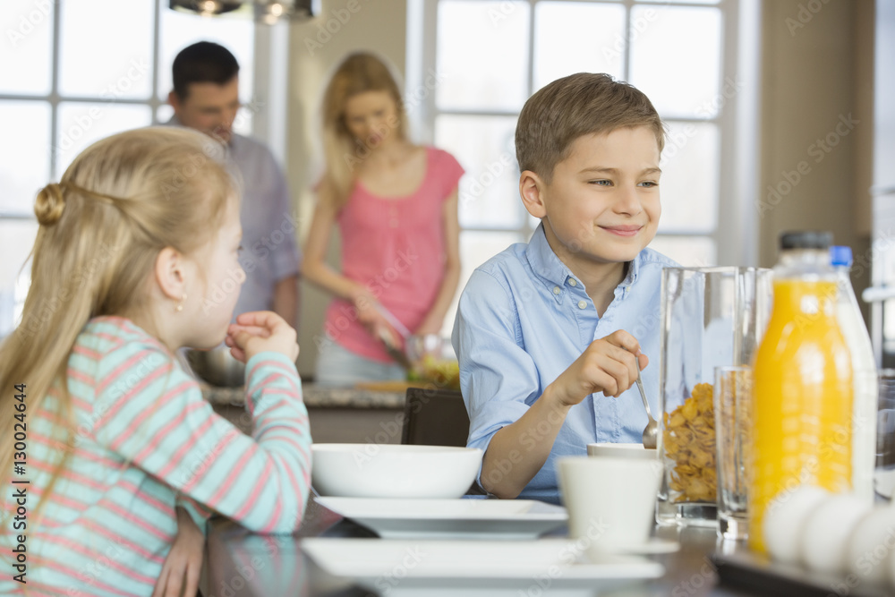 Siblings having breakfast at table with parents cooking in background Siblings having breakfast at table with parents cooking in background