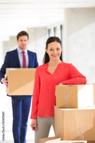 Portrait of confident businesswoman standing by stacked boxes with male colleague in background at office © moodboard