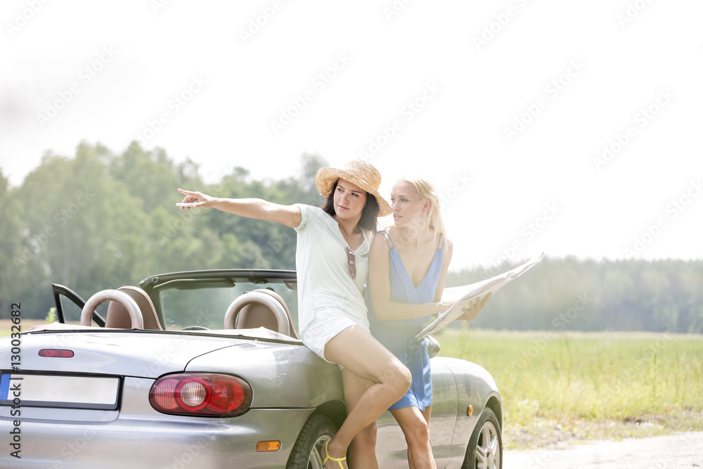 Woman showing something to female friend with map while leaning on convertible
