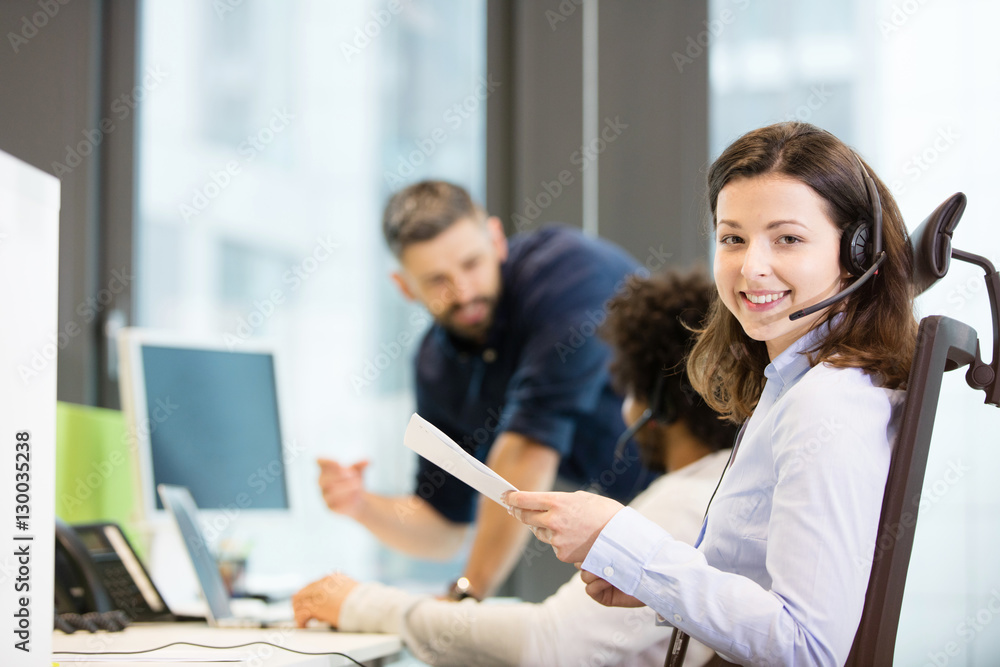 Portrait of smiling customer service representative holding documents with colleagues in background at office Portrait of smiling customer service representative holding documents with colleagues in background at office