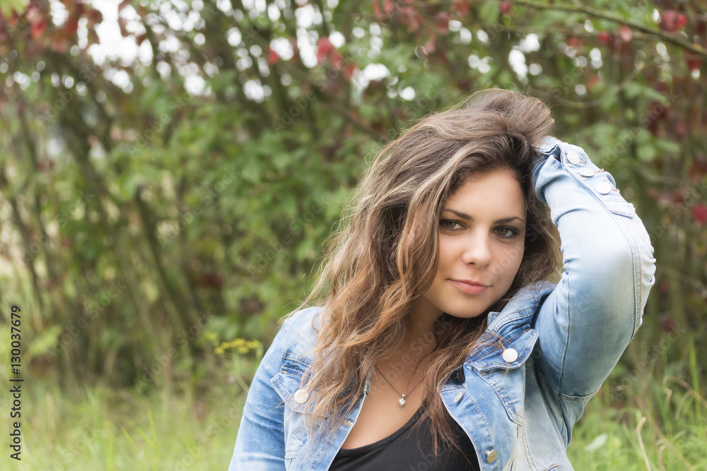 Long haired teenage girl is looking at the camera with hand in her hair and with closed mouth outdoors Long haired teenage girl is looking at the camera with hand in her hair and with closed mouth outdoors