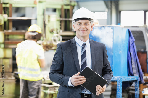 Portrait of confident mature businessman holding digital tablet with worker in background at factory