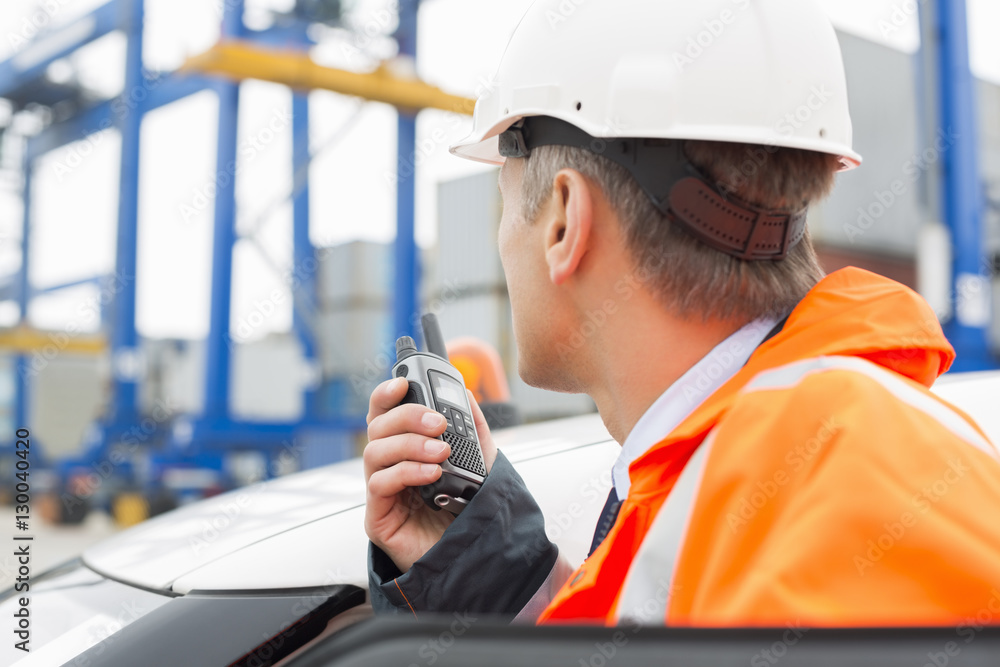 Middle-aged man using walkie-talkie while standing beside car in shipping yard Middle-aged man using walkie-talkie while standing beside car in shipping yard
