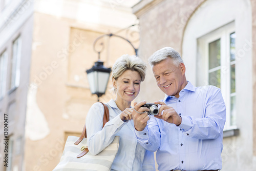 Happy middle-aged couple looking at pictures on digital camera in city