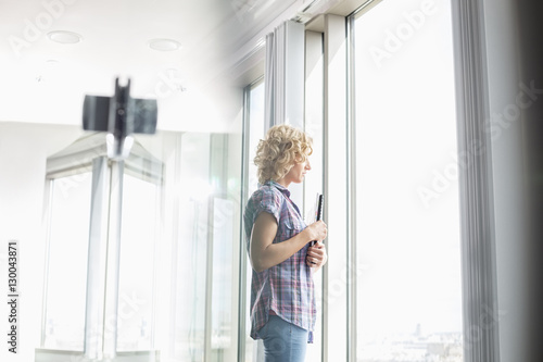 Side view of creative businesswoman holding files while looking through window in office