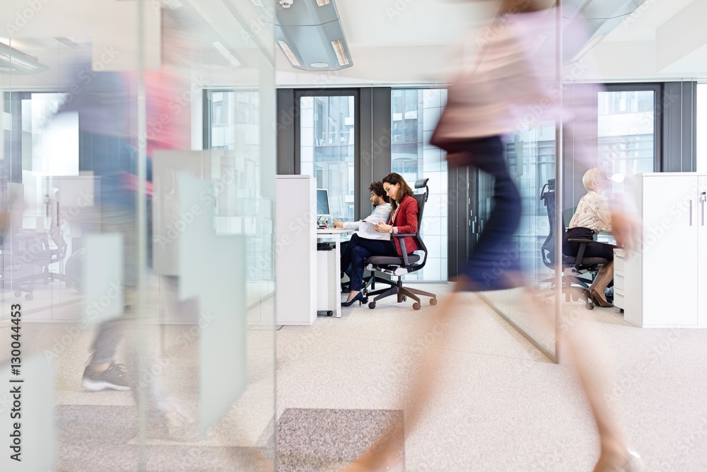 Blurred motion of businesswoman walking with colleagues working in background at office Blurred motion of businesswoman walking with colleagues working in background at office