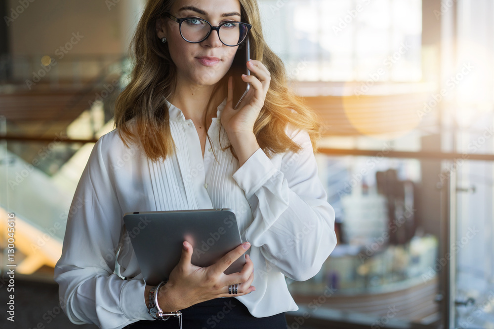 Fototapeta premium Cropped image.Front view. Young business woman in glasses and white shirt, standing in room with modern interior and talking on cellphone while holding tablet computer in her hand. Girl using gadget.