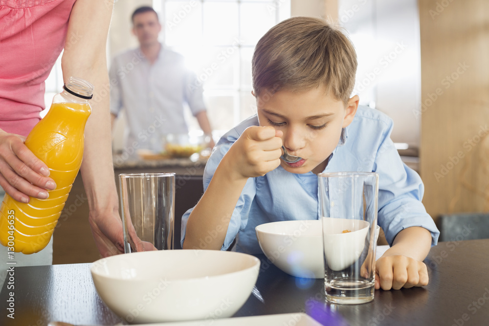 Midsection of woman with juice bottle standing by son having breakfast with man in background Midsection of woman with juice bottle standing by son having breakfast with man in background