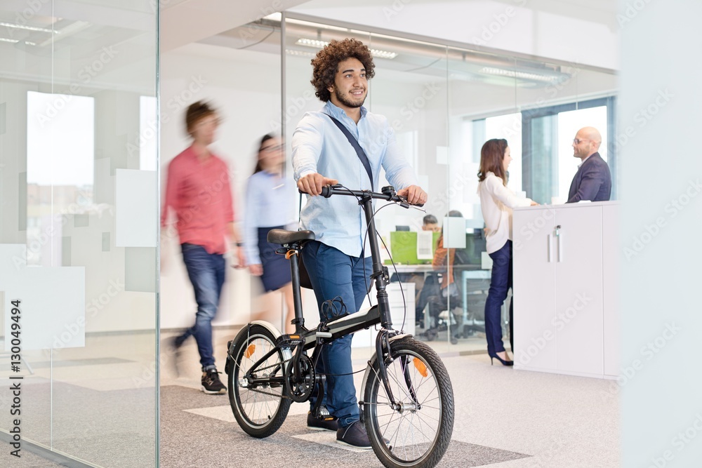 Young businessman walking with bicycle while colleagues in background at office Young businessman walking with bicycle while colleagues in background at office