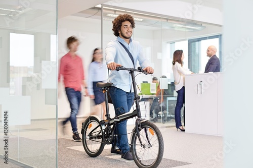 Young businessman walking with bicycle while colleagues in background at office © moodboard
