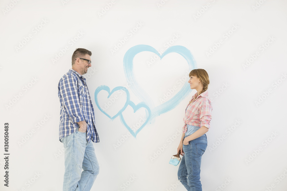 Mid-adult couple looking at each other with painted heart on wall Mid-adult couple looking at each other with painted heart on wall