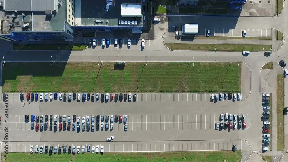 Aerial view of the a capacious parking with different vehicles. Large industrial buildings and green areas are located near the parking lot. Aerial view of the a capacious parking with different vehicles. Large industrial buildings and green areas are located near the parking lot.