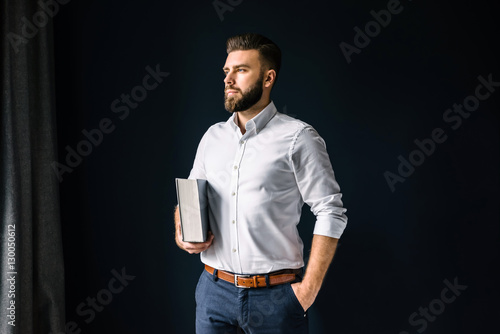 Photos Young bearded businessman, dressed in a white shirt standing near the window on the background of a dark wall and looking out the window