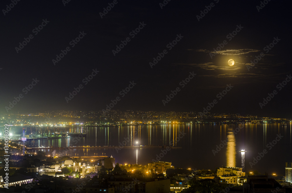 Night view of the city of Baku - the capital of the Republic of Azerbaijan with full moon in soft clouds over dark water with reflections on it