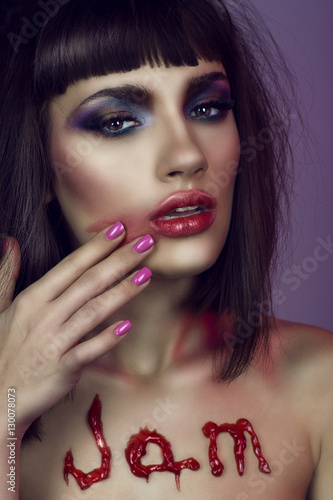 Portrait of young gorgeous woman with bright colourful provocative make-up and dark shaggy hair spreading red lipstick over her face. The word JAM written on her naked chest. Close up