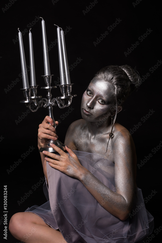 Beautiful serious and sad young topless woman with silver body art and nice hairstyle wearing gray veil skirt sitting on the floor and keeping candlestick with candles in one hand