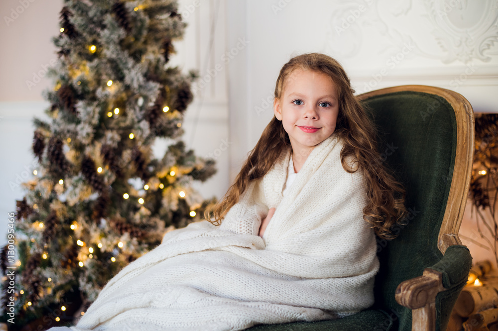 Cute little girl sits by the christmas tree, wrapped in a warm blanket at home Cute little girl sits by the christmas tree, wrapped in a warm blanket at home