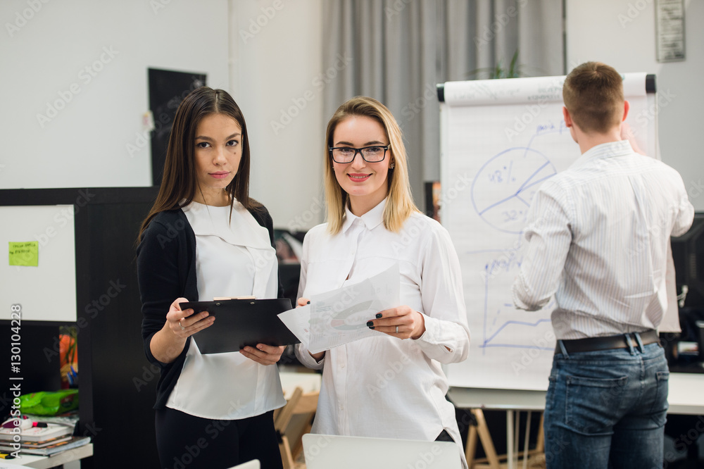 Two beautiful office workers having conversation while man colleague drawing business strategy on flip chart. Two beautiful office workers having conversation while man colleague drawing business strategy on flip chart.