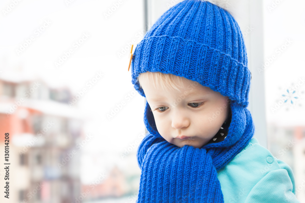 Sick child in warm clothes with sad look during winter holidays in house opposite window, wishing to go walking Sick child in warm clothes with sad look during winter holidays in house opposite window, wishing to go walking