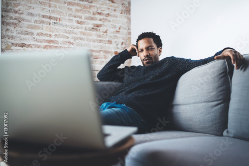 Young African confident businessman sitting on sofa at his modern coworking office and making video call via laptop.Concept of serious business people.Blurred background. photo