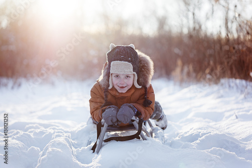 Cute little funny child boy in warm winter clothes having fun on snow sledge, outdoors during snowfall.