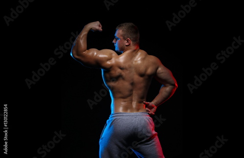 male bodybuilder athlete with naked torso posing against a black background, in red and blue light