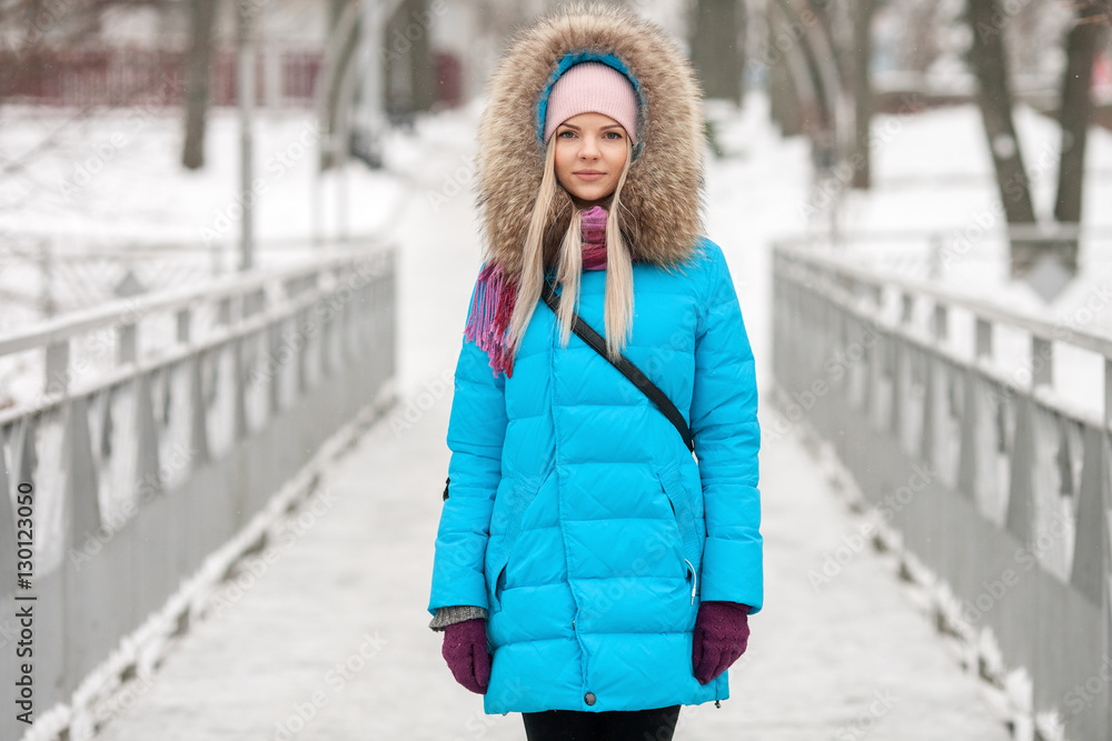 Young adorable blond woman wearing blue hooded coat strolling in snowy winter city park bridge. Nature cold season freshness concept. Young adorable blond woman wearing blue hooded coat strolling in snowy winter city park bridge. Nature cold season freshness concept.