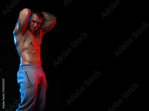 male bodybuilder athlete with naked torso posing against a black background, in red and blue light