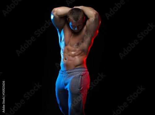 male bodybuilder athlete with naked torso posing against a black background, in red and blue light