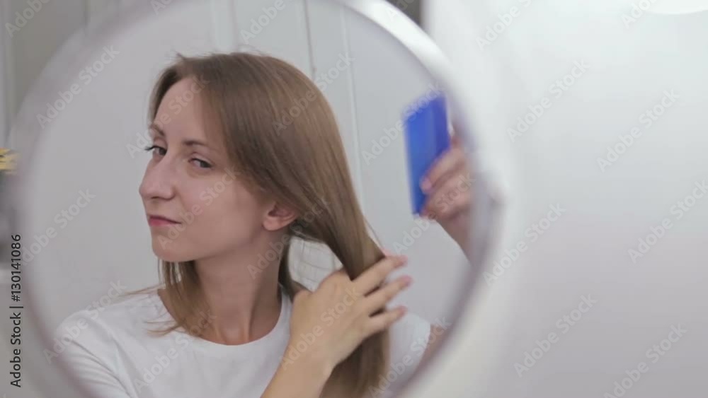 Young pretty woman combing her hair in front of the mirror. White interior, makeup room Young pretty woman combing her hair in front of the mirror. White interior, makeup room