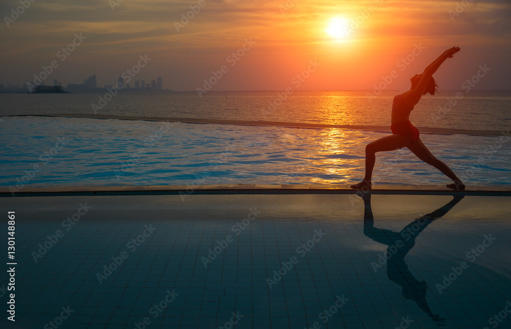 Silhouette young woman practicing yoga on swimming pool and the beach at sunset