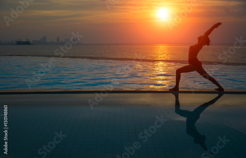 Silhouette young woman practicing yoga on swimming pool and the beach at sunset