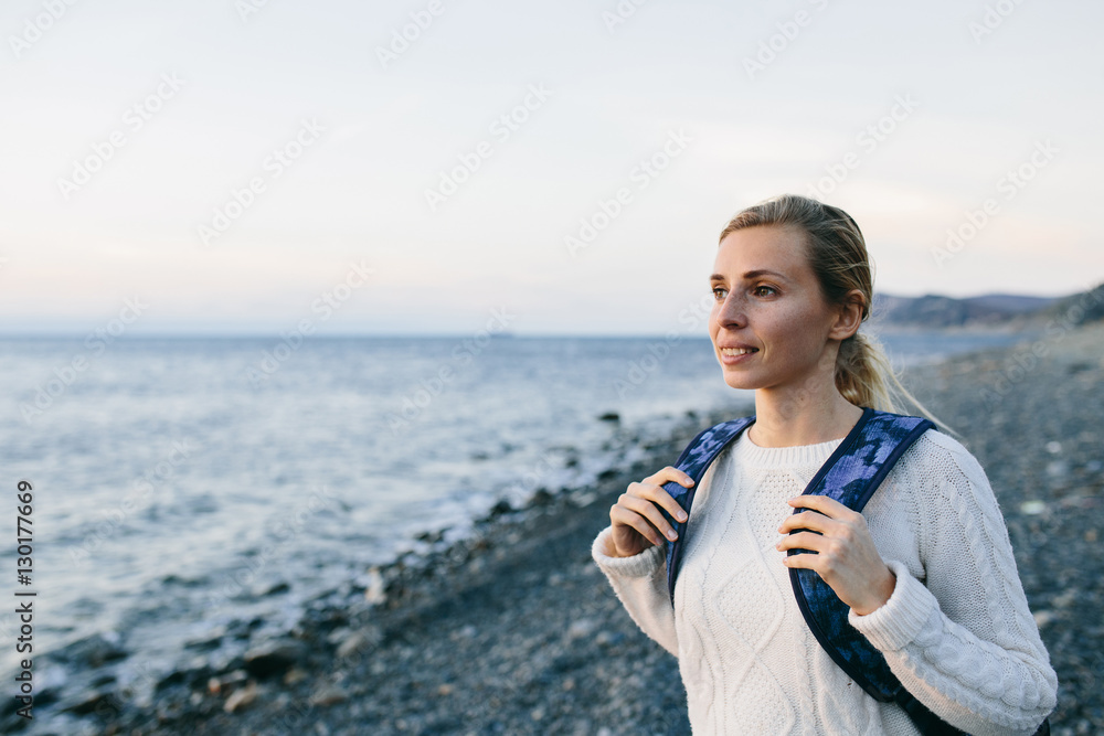 smiling young woman traveler in a white clothing standing on the shore and looks at sea