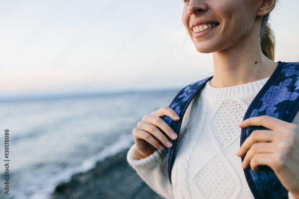 Close up of smiling young woman traveler in a white clothing standing on the shore and looks at sea