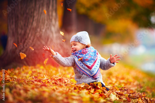 beautiful happy baby girl having fun in autumn park, among fallen leaves