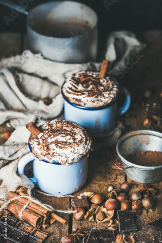 Hot chocolate in blue mugs with whipped cream and cinnamon sticks, spices, nuts and cocoa powder on rustic wooden background, selective focus, vertical composition